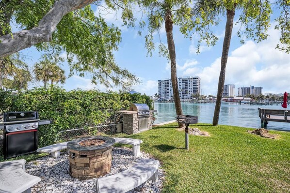 An outdoor patio with a grill, a fire pit, and a view of a body of water and buildings in the background.