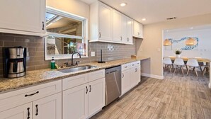 A modern kitchen with white cabinets, a granite countertop, a built-in dishwasher, and a dining area with a wooden table and chairs. - Sunny Home & Yard, Minutes to Beach (Seminole)