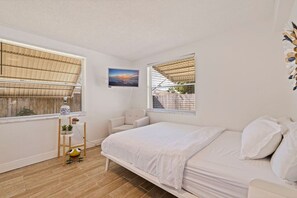 Ein Schlafzimmer mit einem Bett, zwei Fenstern mit Jalousien, einem Sessel, einem kleinen Tisch mit Lampe und einem Bild an der Wand.