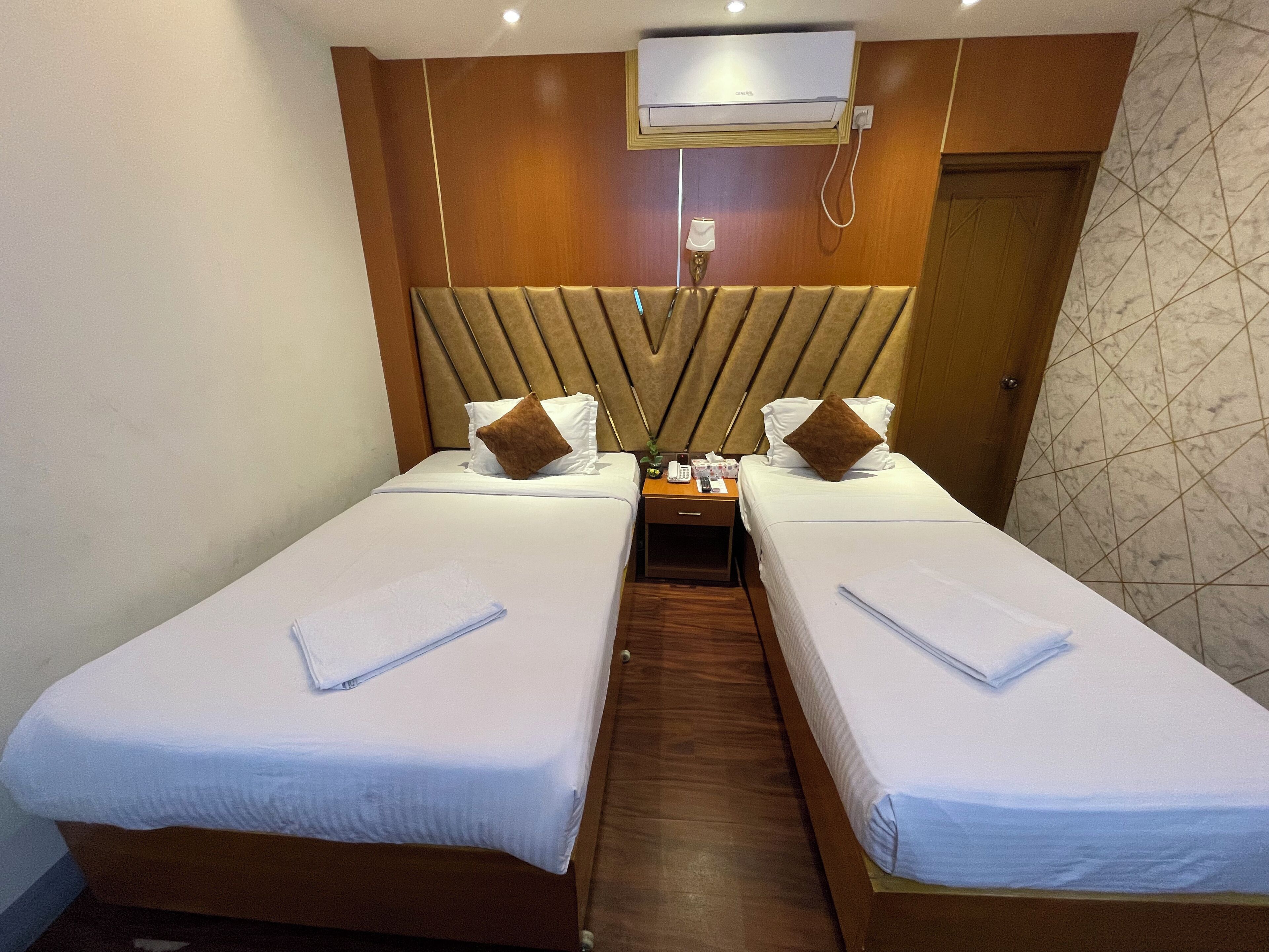 A hotel room with two single beds, a bedside table with a phone and a lamp, and a wall-mounted air conditioner.