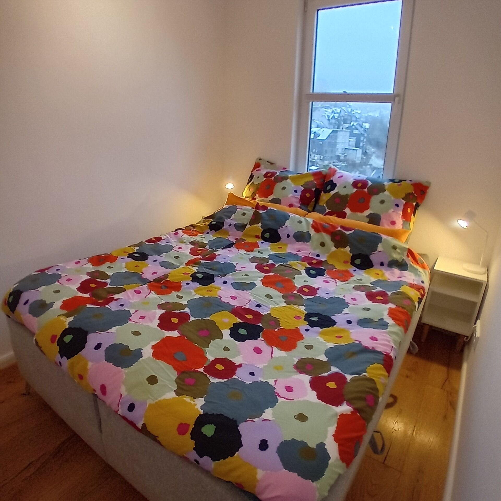 A bed with a colorful floral bedspread and matching pillows, a bedside table with a lamp, and a window with a view of buildings.