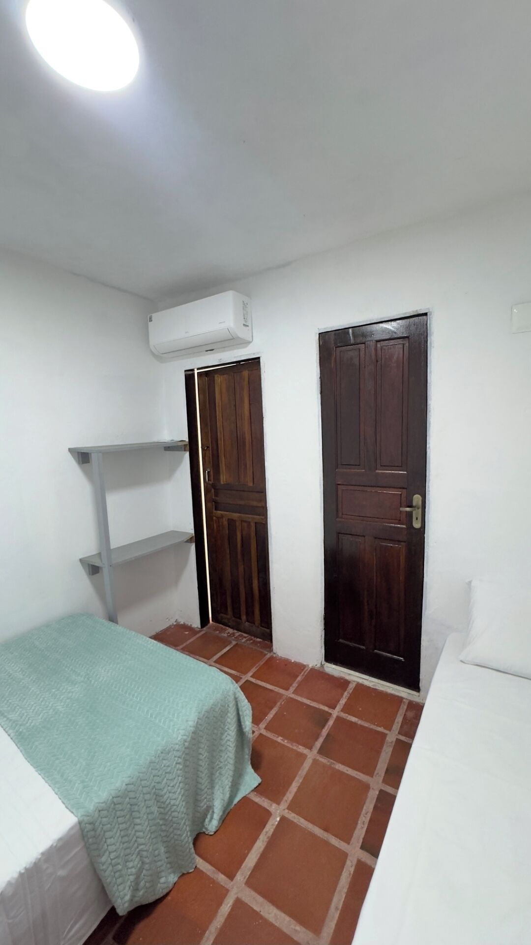 Een kleine, eenvoudige kamer met een bed, een plank, een deur en een airconditioner.