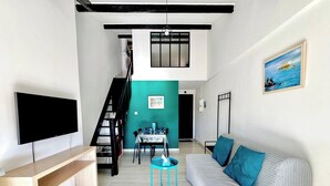 A modern living room with a flat-screen TV, a sofa with blue cushions, a dining area with a table and chairs, and a staircase leading to an upper level.