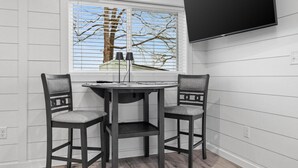 A room with a table and two chairs, a window with blinds, and a mounted TV. - Unique Shipping Container Stay - Walk to Branson Landing! (Branson)