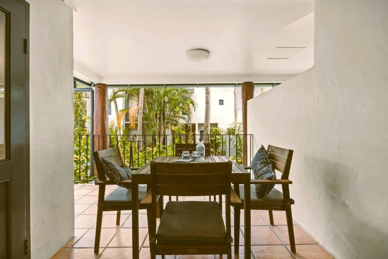 A balcony with a wooden table and chairs, a view of palm trees, and a building in the background.