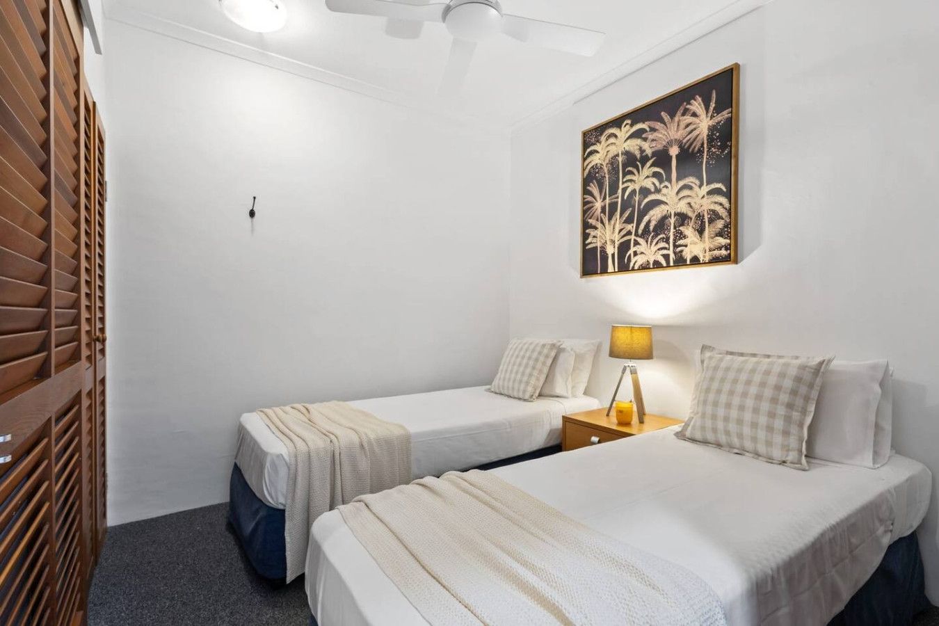 Una camera d'albergo con due letti, un armadio in legno, una ventola a soffitto e un'opera d'arte appesa al muro.