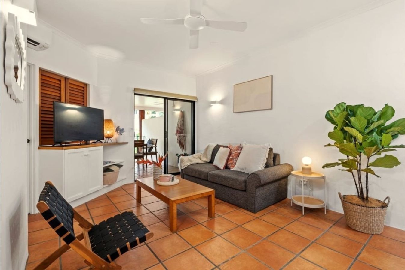 Una sala de estar con un sofá, una mesa de centro y una televisión. Hay una planta y una mesita de noche con una lámpara.