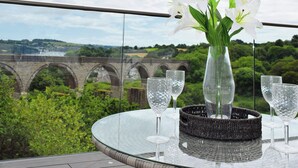 A table with a vase of white flowers and three wine glasses on a glass table with a view of a bridge and greenery in the background.