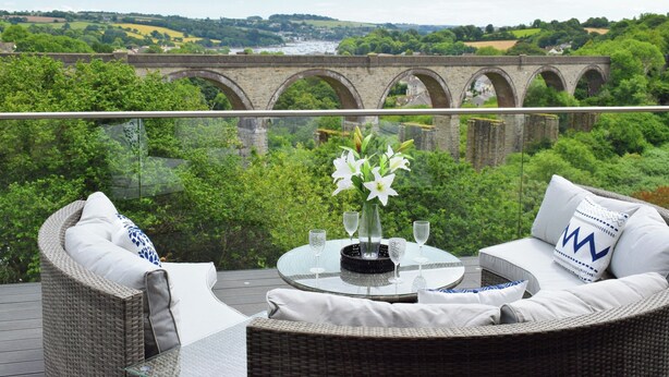 A balcony with wicker furniture, a glass table with a vase of flowers, and a view of a stone bridge over a river.