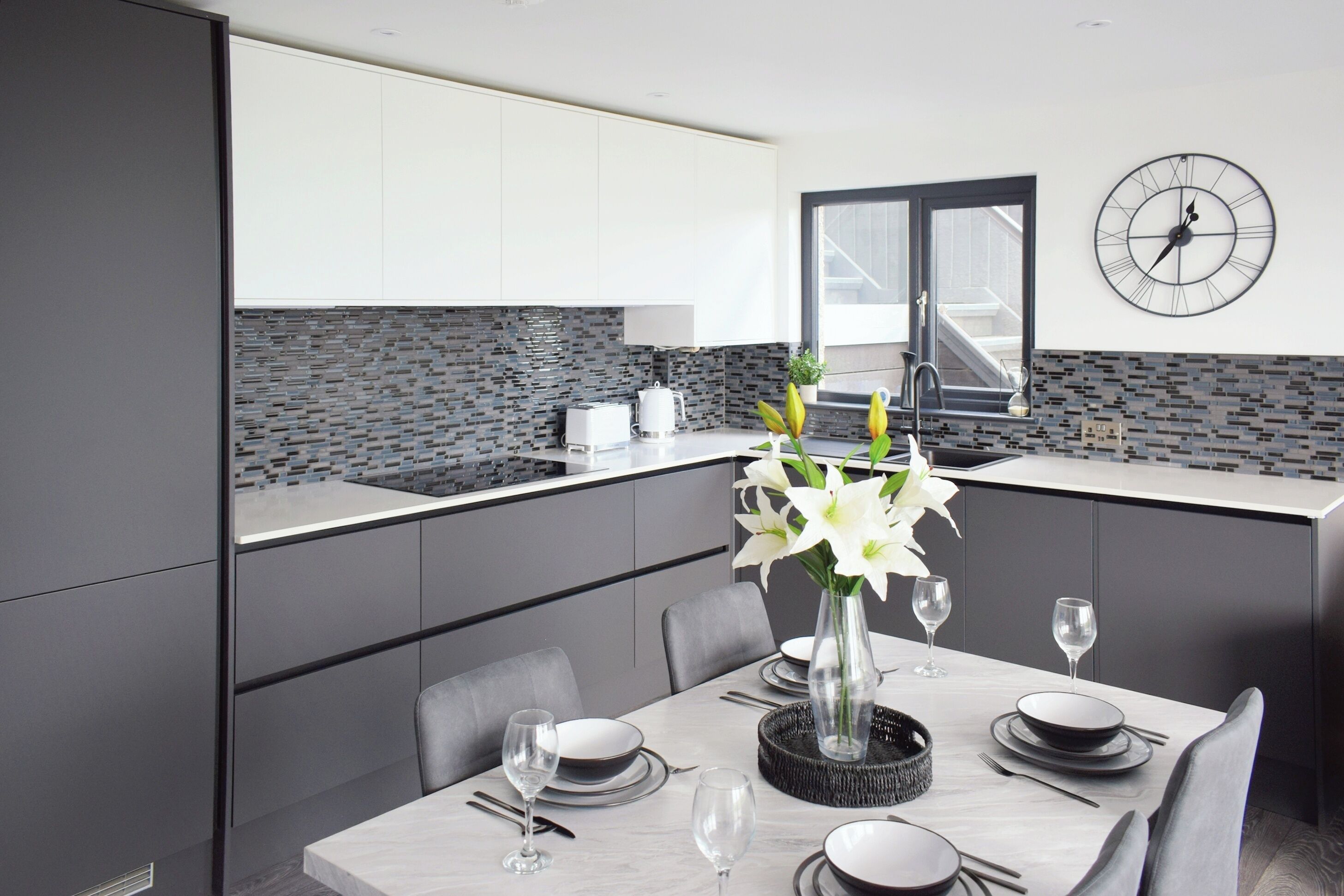 A modern kitchen with grey cabinets, a tiled backsplash, and a dining area set for six with a vase of flowers.
