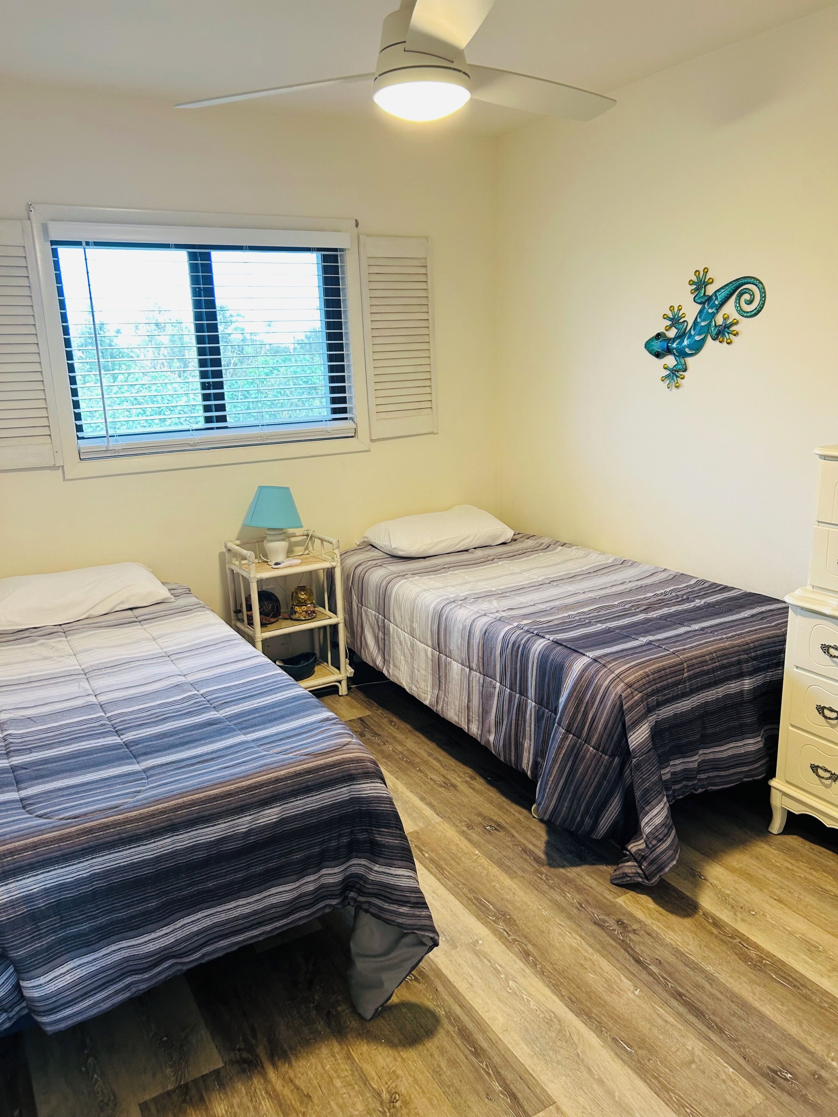 Une chambre avec deux lits, une table de chevet, une commode, un ventilateur de plafond et une fenêtre avec des stores.