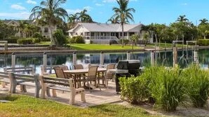 A lakeside view with a patio area, lounge chairs, and a house in the background.