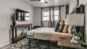 A bedroom with a bed, a desk, a TV, a ceiling fan, and a window with curtains.