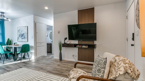 A modern living room with a flat-screen TV mounted on a wooden panel, a dining area with green chairs, and a kitchen visible through an open door.
