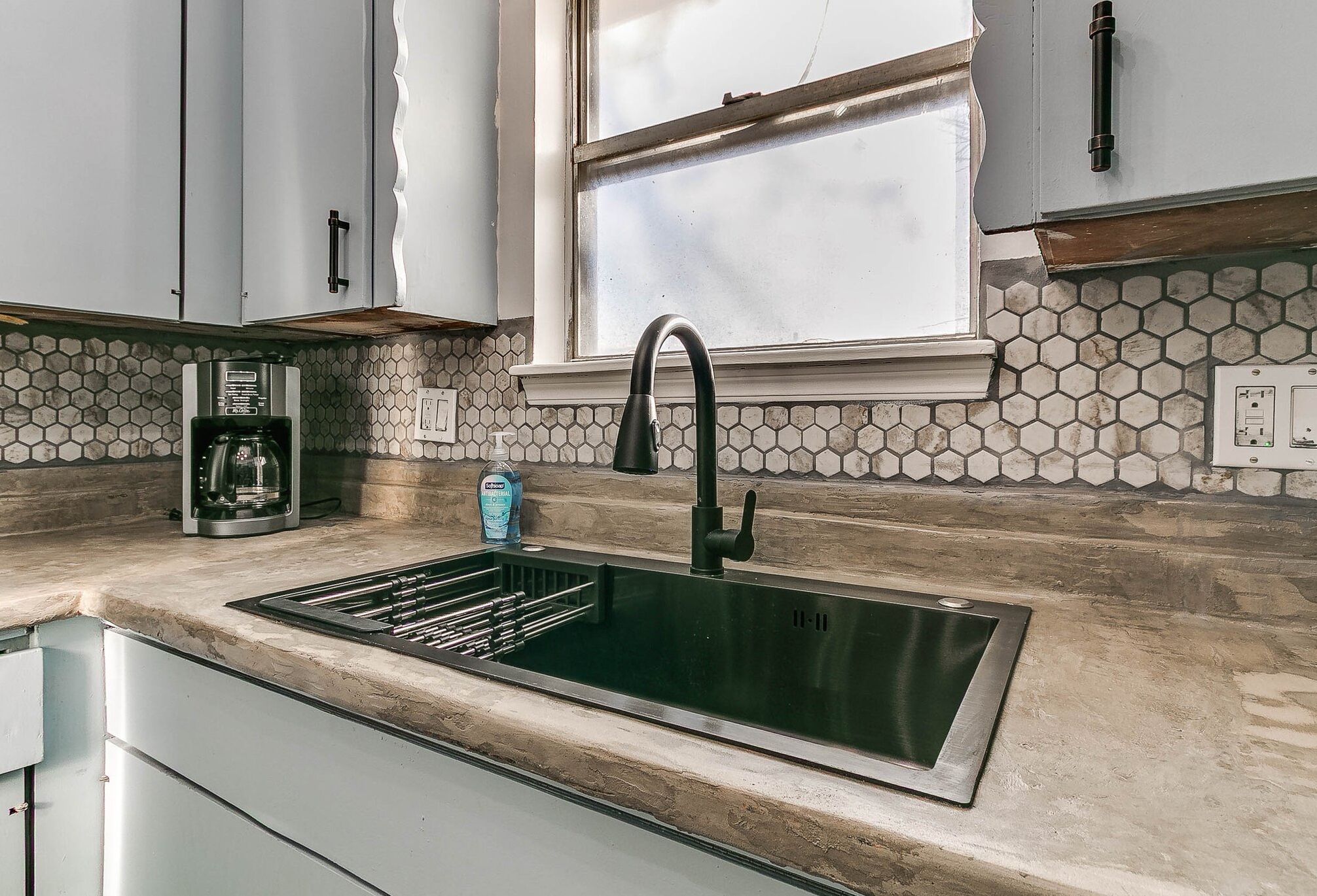 Una cocina con un lavamanos negro, una cafetera y una ventana sobre el lavamanos.