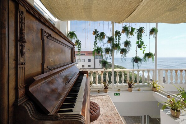 Un piano en un balcón con vistas al mar, plantas colgantes y una edificación a la distancia.