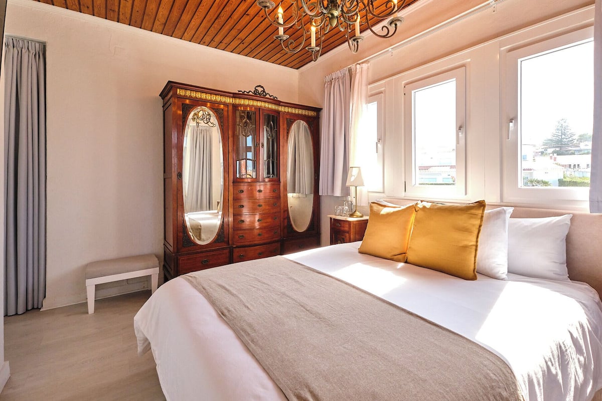 Ein Schlafzimmer mit einem großen Bett, einer Holzkommode, einer Bank und zwei Fenstern.