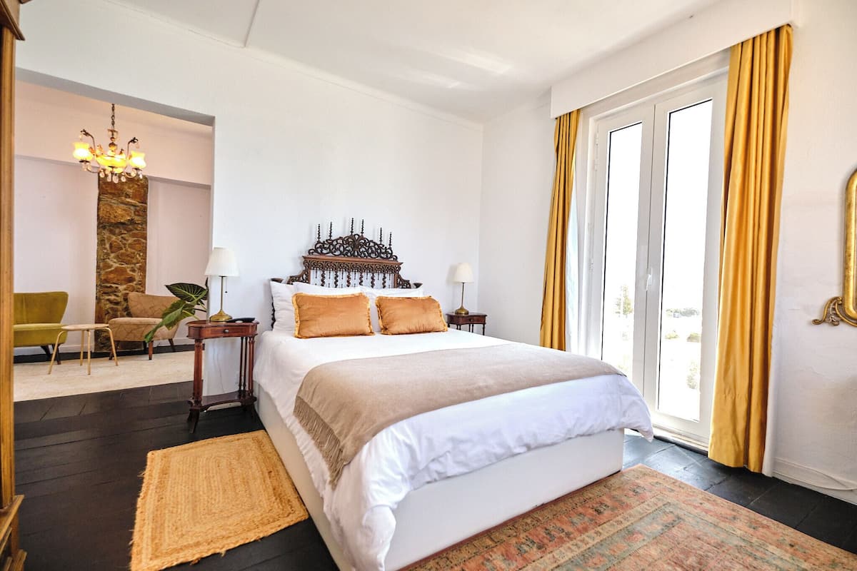 Ein Hotelzimmer mit einem großen Bett, einer Kristallleuchtre, einer Steinwand, einem Sessel und Blick durch eine Schiebetür.