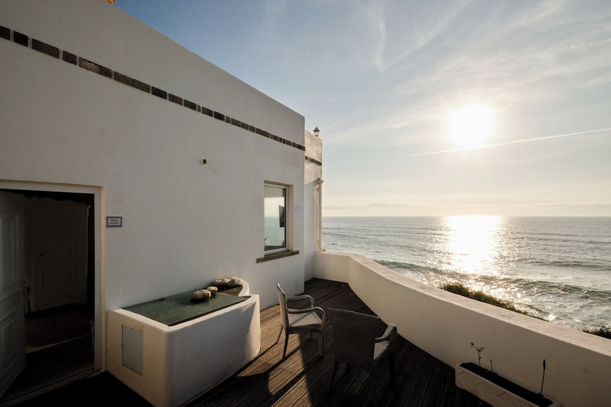 Ein Balkon mit Blick auf das Meer, eine kleine Sitzecke und eine Tür zu einem Innenraum.