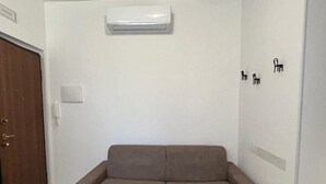 Una habitación con un sofá color beige, una puerta de madera y una pared blanca con un termostato y un interruptor de luz.