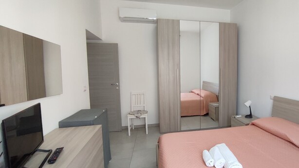 Un dormitorio moderno con una cama, un escritorio con televisor y control remoto, una silla, una mesita de noche con lámpara y un armario con espejo.