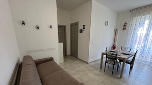 Una sala de estar con un sofá marrón, una mesa de comedor con sillas, una pequeña cocina y una puerta que da a otra habitación.