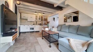 Un salon spacieux avec un canapé gris, une table basse et un téléviseur. Au fond de la pièce, on aperçoit une cuisine avec des meubles blancs et un dosseret carrelé.