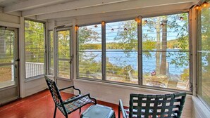 A sunroom with large windows overlooking a lake, featuring two chairs and a small table. - Stonedam Cottage on the shores of Lake Winni (Meredith)