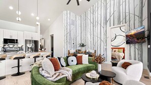Un salon moderne avec un canapé d’angle vert, des fauteuils blancs et une table basse. Un téléviseur à écran plat est fixé au mur, et il y a un coin cuisine avec des meubles blancs et des appareils en acier inoxydable.
