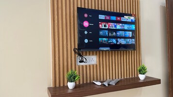 Un televisor de pantalla plana montado en una pared de madera, con una repisa debajo que muestra dos plantas en macetas y un control remoto.