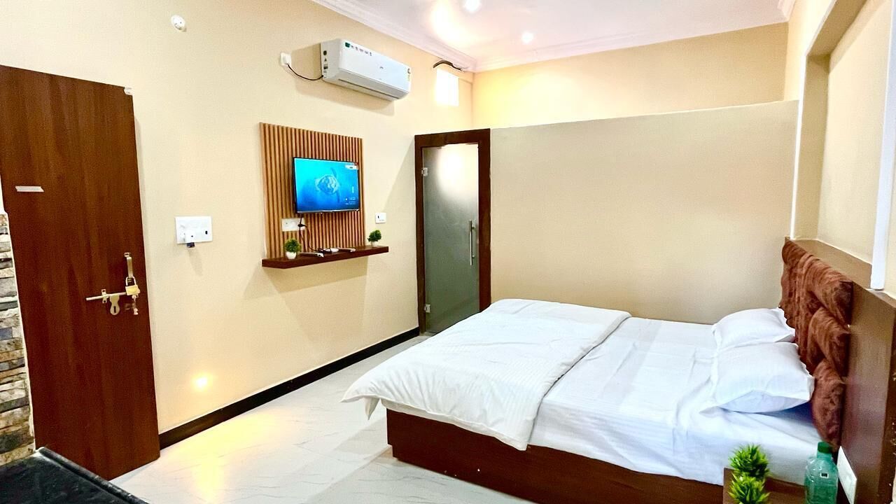 A hotel room with a bed, a TV mounted on the wall, a wooden door, and a small shelf with a plant.