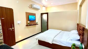 A hotel room with a bed, a TV mounted on the wall, a wooden door, and a small shelf with a plant.