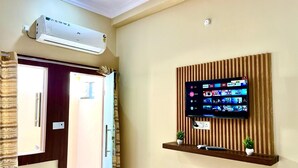 A hotel room with a mounted TV displaying various streaming services, a wall-mounted air conditioner, and a wooden shelf with a plant. - Raghav ji home stay (Orchha)