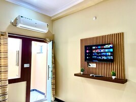 A hotel room with a mounted TV displaying various streaming services, a wall-mounted air conditioner, and a wooden shelf with a plant.
