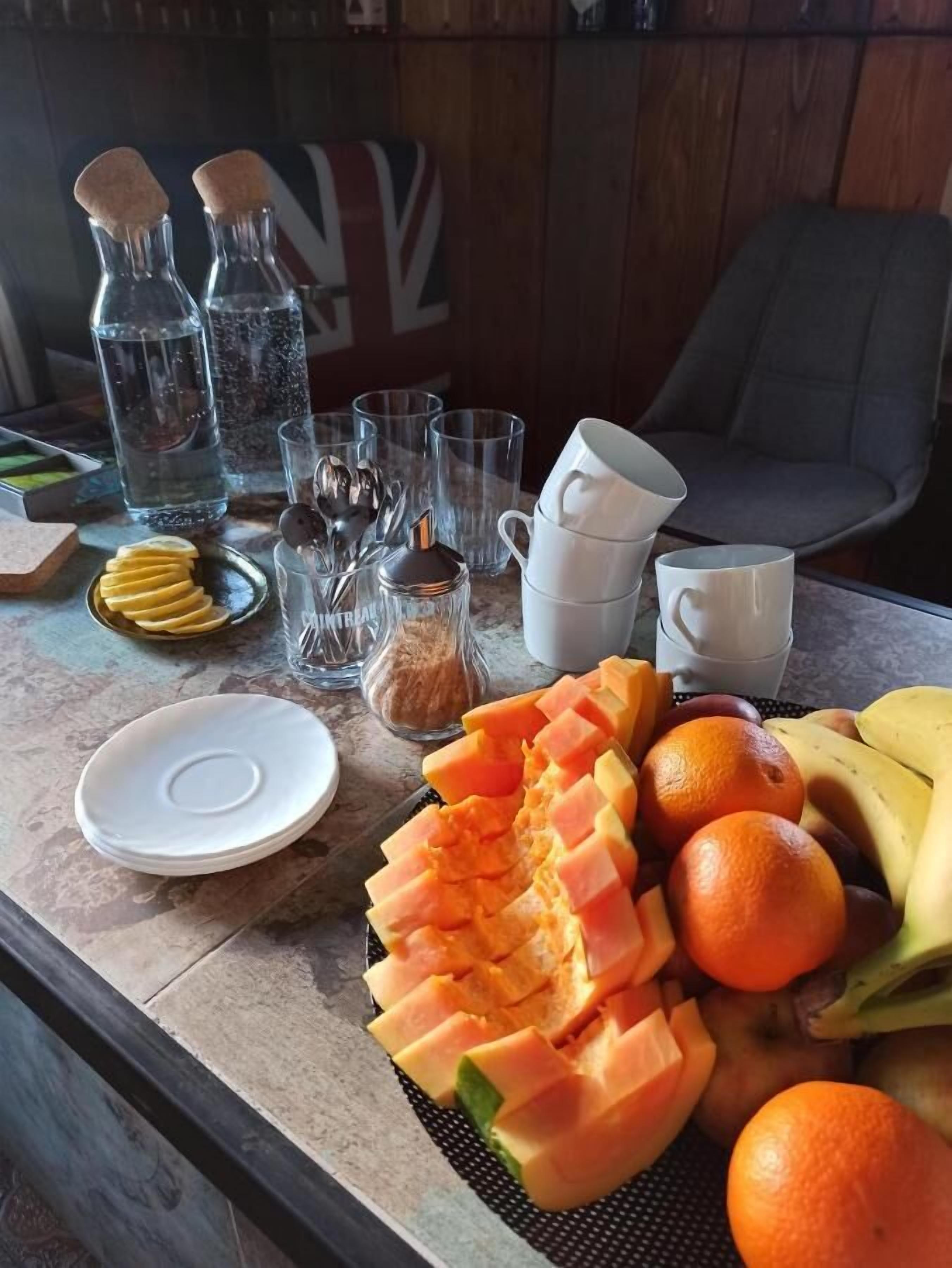 A table with a variety of fruits, including sliced melon, oranges, and bananas, along with several glasses and a bottle.