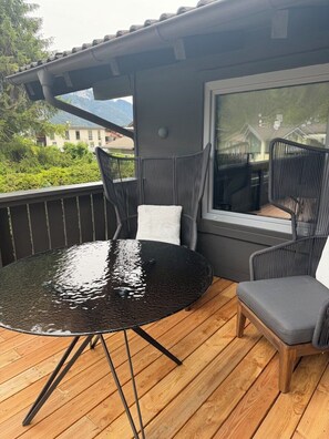 A deck with a round table and two chairs, a window with a view of a house and mountains, and a wooden floor.