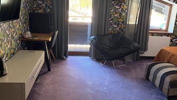 Ein Zimmer mit Flachbildfernseher, Schreibtisch mit Computer, einer Sitzecke und einem Fenster mit Vorhängen.