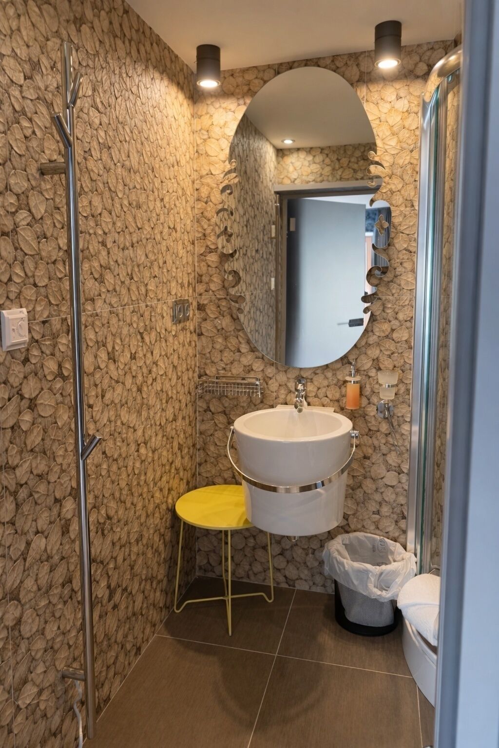 A bathroom with a stone wall, a white sink, a yellow stool, and a glass shower enclosure.