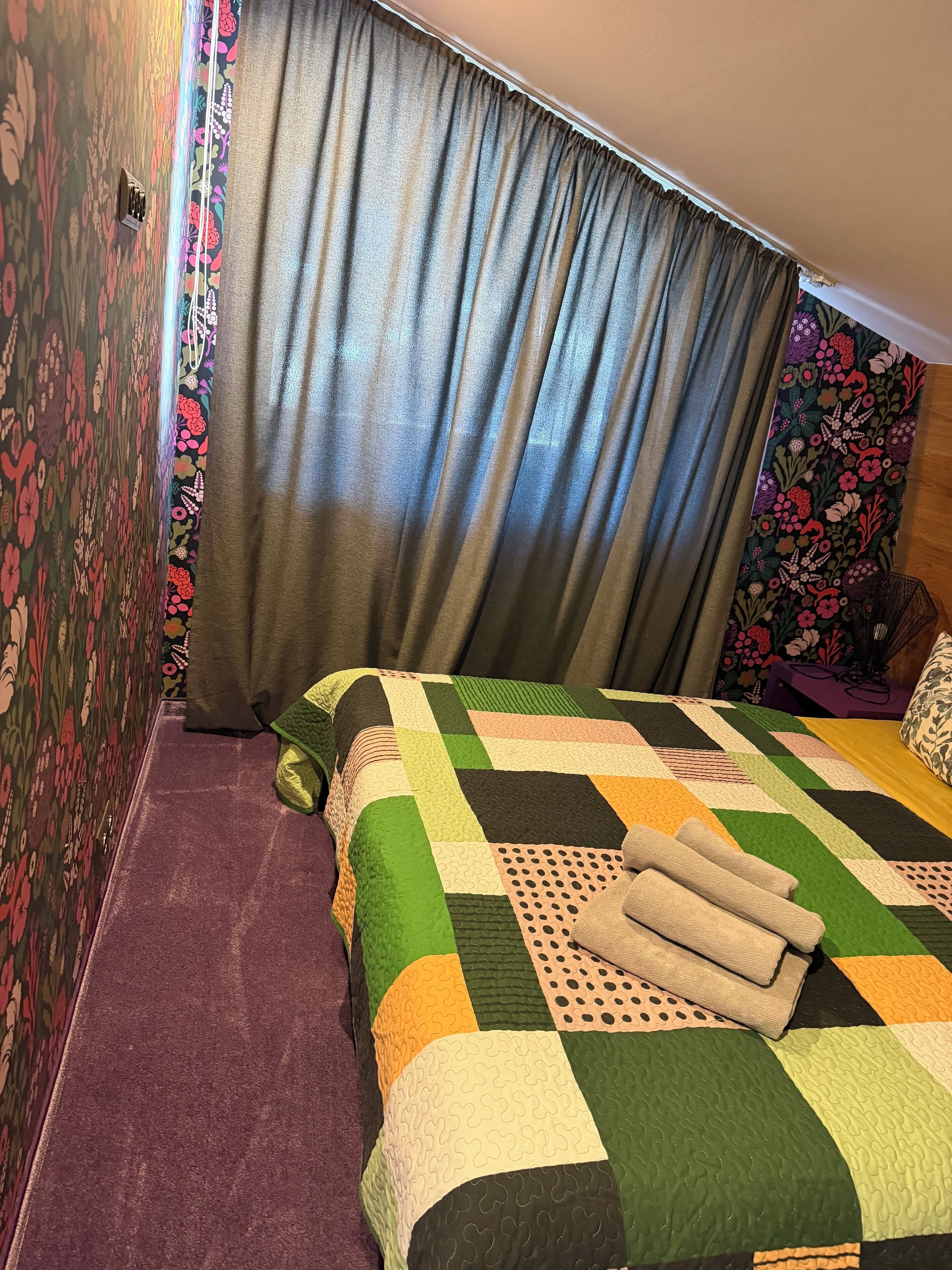A hotel room with a bed, a purple carpet, floral wallpaper, and a window with curtains.