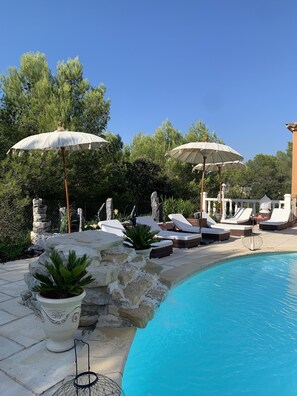 A poolside area with lounge chairs, umbrellas, and a stone wall. - LES HUPPES Guest rooms nestled in the heart of the garrigue with swimming pool (Nîmes)