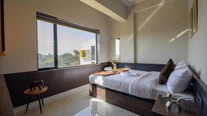 A modern bedroom with a large window, a bed with white linens, a wooden side table, and a small round table with a chair. - Saavi Suites (Daman)