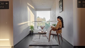 A modern interior with a person sitting on a chair, a potted plant, a small round table, and a view through a window. - Saavi Suites (Daman)