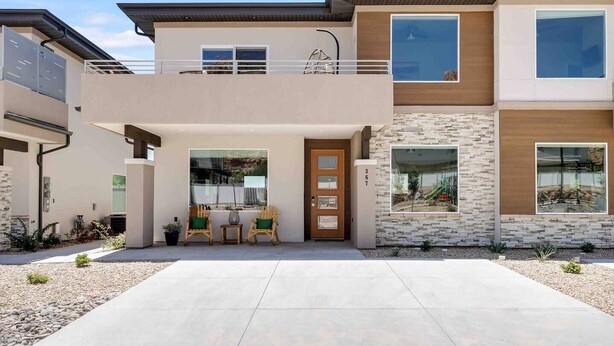 A modern two-story house with a balcony, a front door, and a small patio area with chairs and a table.