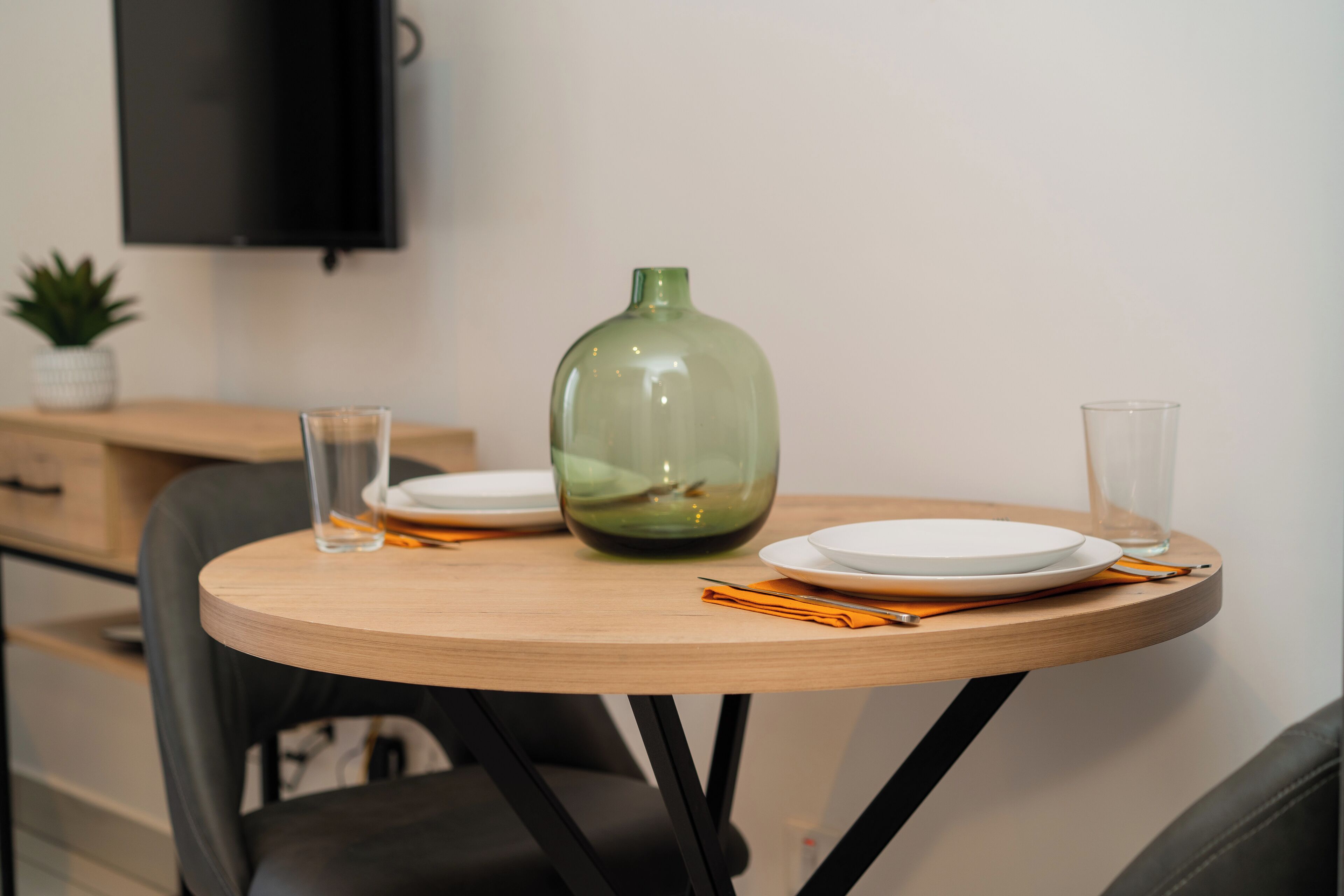 A round wooden table with a green vase, two white plates, and two glasses.