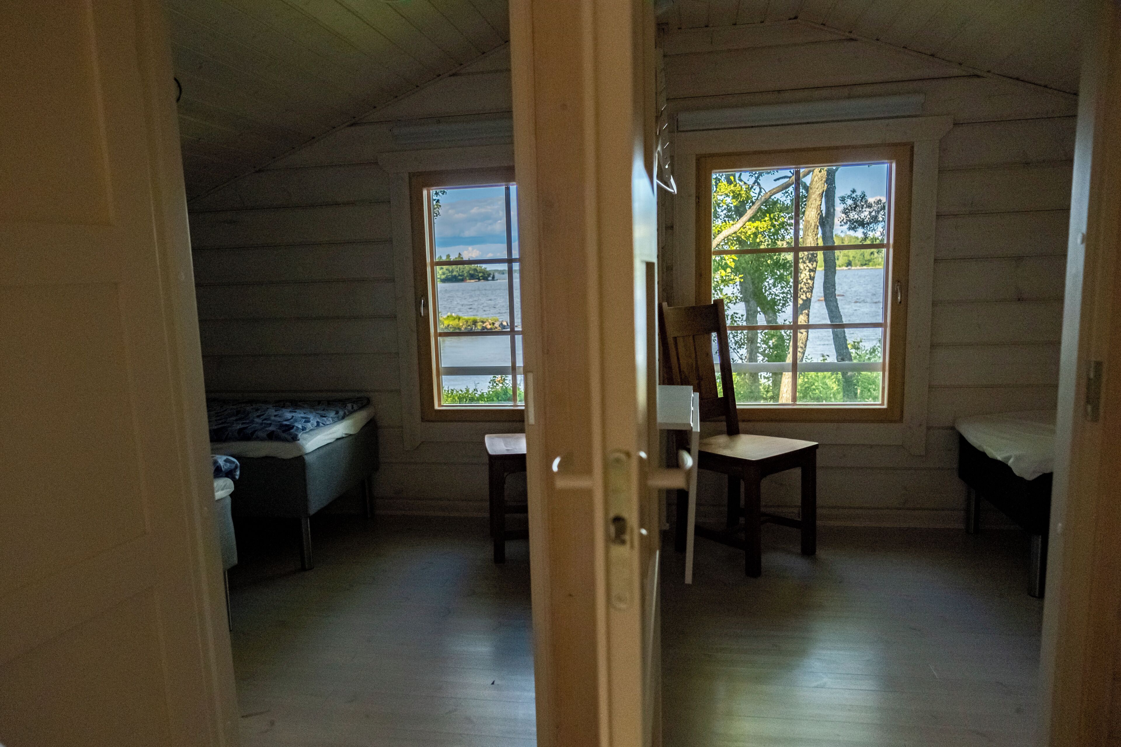 Une cabane avec deux lits, une table et une chaise. Il y a des fenêtres donnant sur les arbres et l’eau.