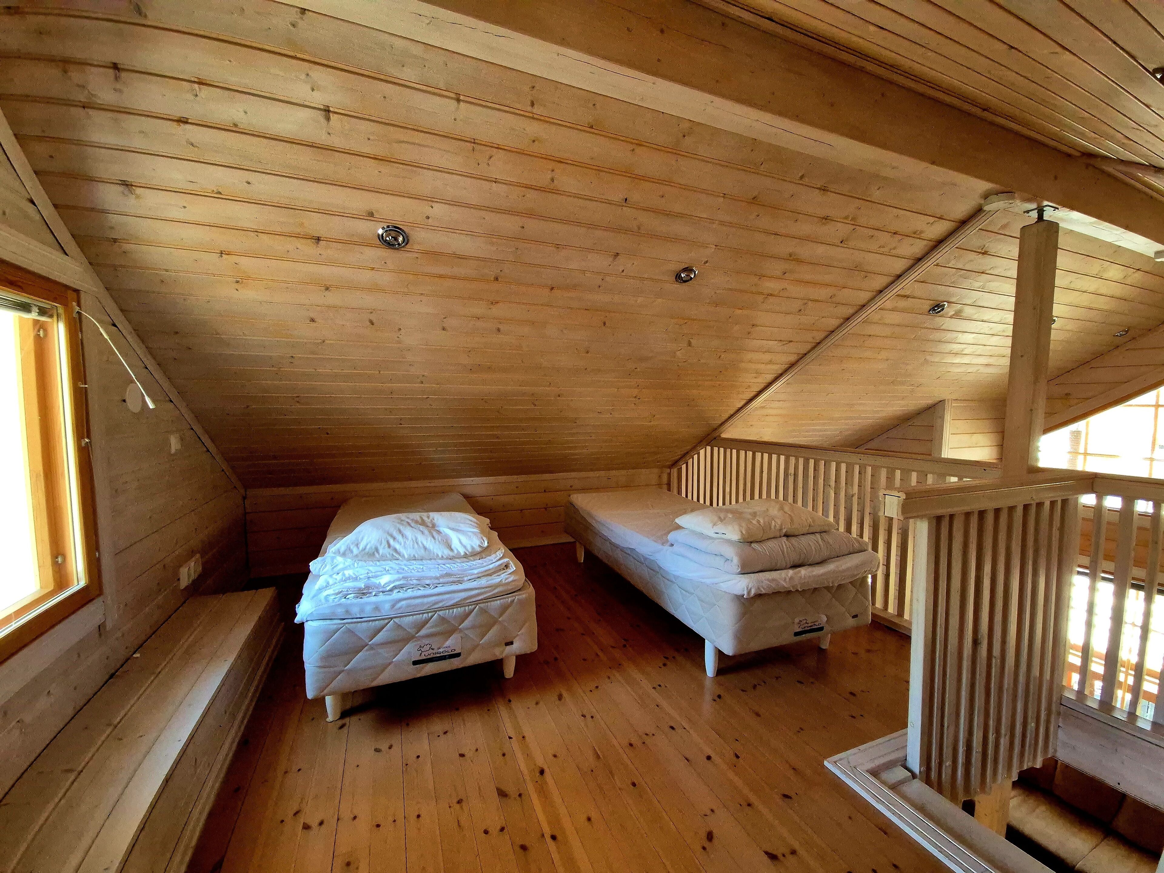A small, wooden attic bedroom with two beds, a window, and a wooden railing.