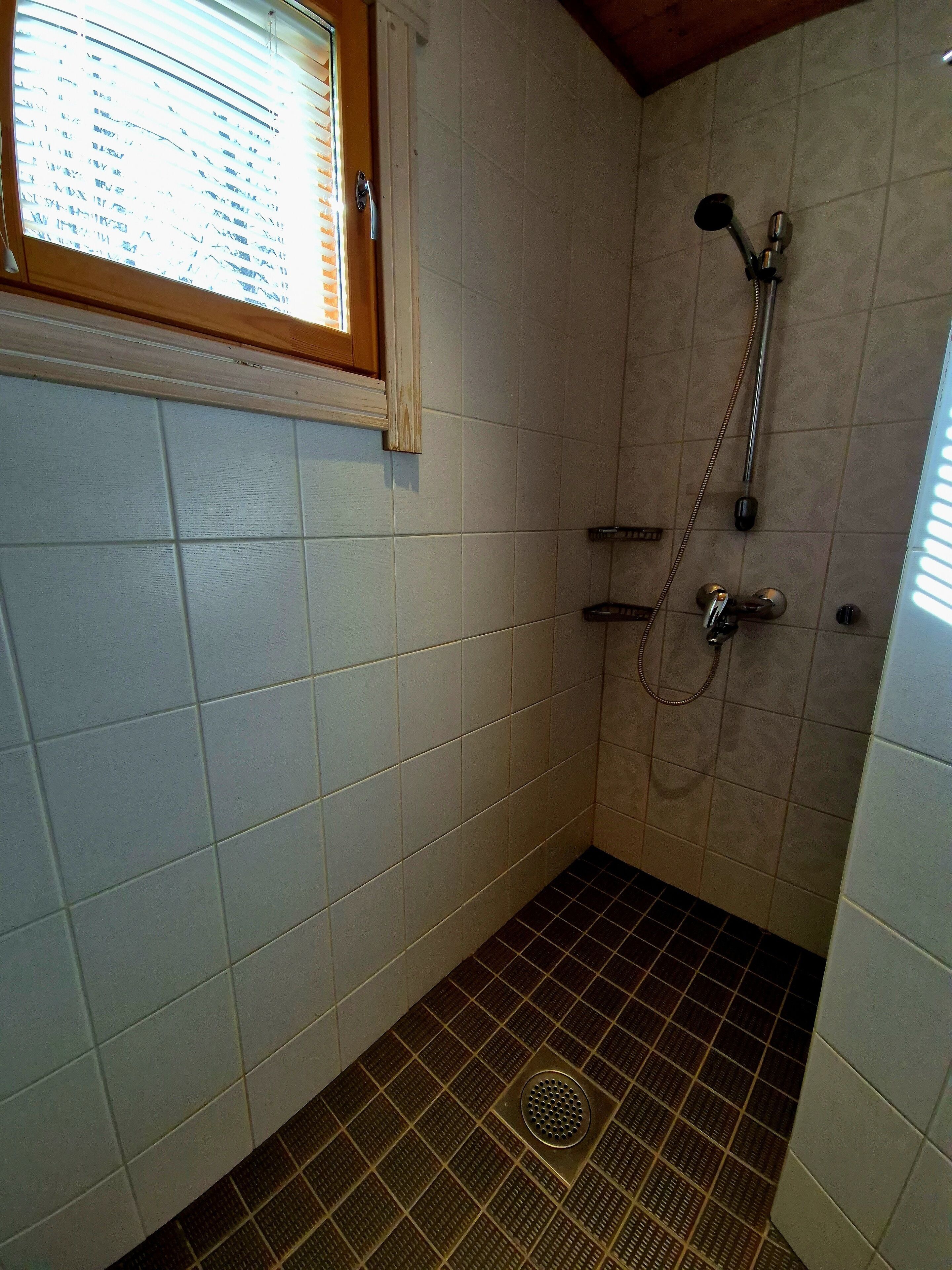 A small bathroom with white tiled walls, a shower with a handheld showerhead, and a window with blinds.