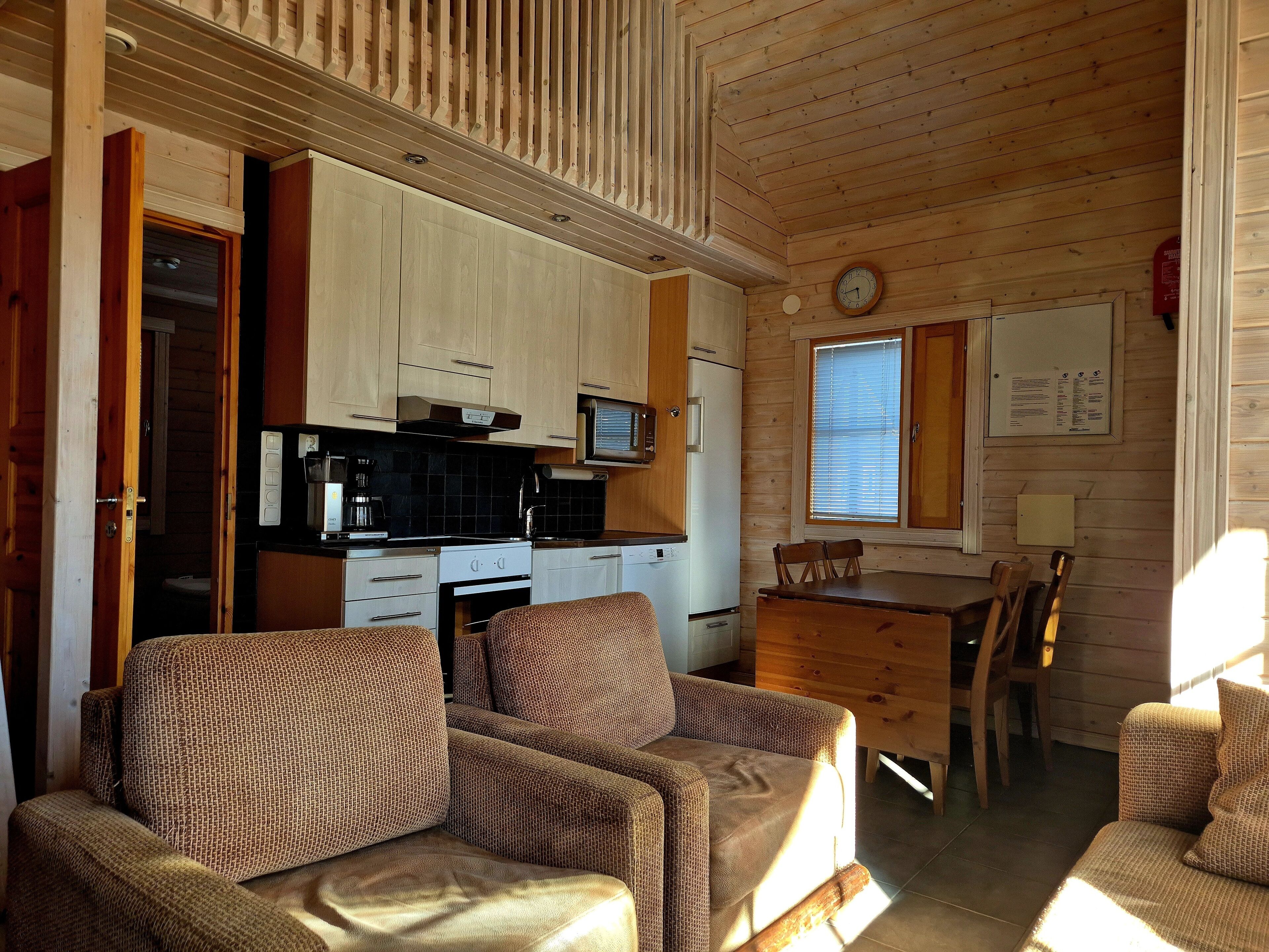 Interior acogedor de una cabaña con kitchenette, paredes de madera, zona de comedor con mesa y sillas, y un sofá.