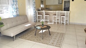 A living room with a sofa, a coffee table, and a patterned rug. There is a kitchen area with white cabinets and a dining area with white chairs.