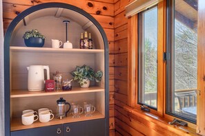 Interior de una cabaña de madera con un estante curvo que contiene un hervidor, tazas, una cafetera y botellas.